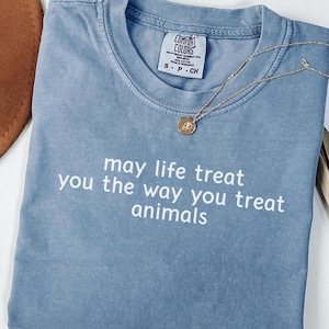 May include: A blue short-sleeved t-shirt with a crew neckline and the white text "may life treat you the way you treat animals" printed on the front. The shirt features a Comfort Colors brand label at the inner neck with size S P CH.