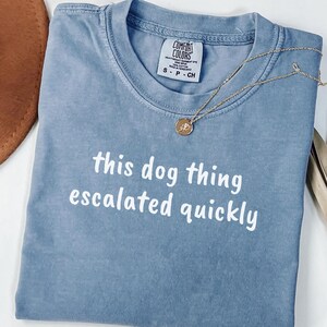 May include: A blue short-sleeved t-shirt with the white text "this dog thing escalated quickly" printed on the front. The shirt features a Comfort Colors brand label at the neckline and is displayed with a gold-toned pendant necklace.