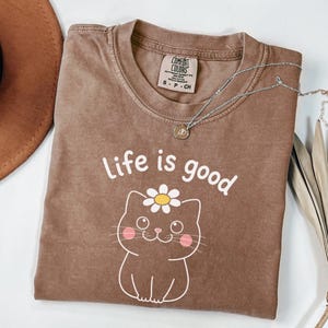 May include: Brown t-shirt with the phrase "Life is good" and a cartoon cat graphic with a daisy. A silver necklace with a pendant is draped across the shirt. A brown hat is visible in the upper left corner. The shirt is labeled Comfort Colors.