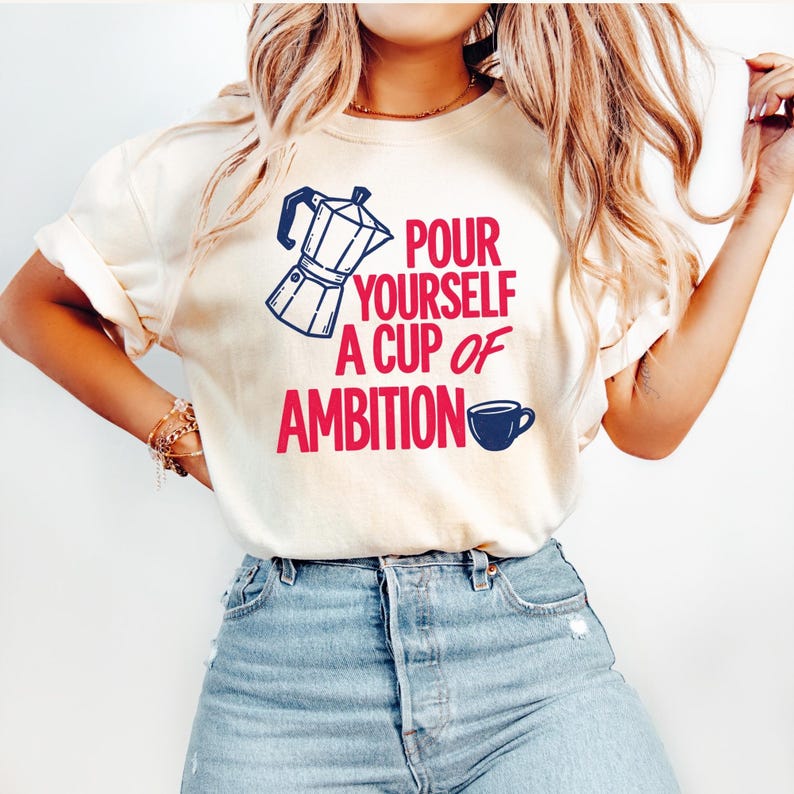 Puede incluir: Una camiseta de color crema con un gr&aacute;fico de una cafetera italiana azul oscuro y una peque&ntilde;a taza de caf&eacute; azul oscuro. El texto en rojo dice "POUR YOURSELF A CUP OF AMBITION". La camiseta tiene mangas cortas y cuello redondo, combinada con vaqueros azul claro.