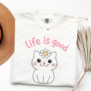 May include: White t-shirt with the phrase "Life is good" in pink, above a cartoon cat wearing a daisy. A gold necklace with a star pendant and a brown hat are also visible.