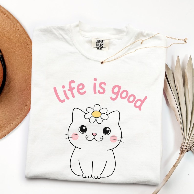 May include: White t-shirt with the phrase "Life is good" in pink, above a cartoon cat wearing a daisy. A gold necklace with a star pendant and a brown hat are also visible.