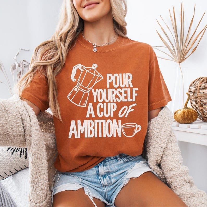 Puede incluir: Una camiseta de manga corta color naranja quemado con un gr&aacute;fico blanco de una cafetera italiana y una taza de caf&eacute;, con el texto "POUR YOURSELF A CUP OF AMBITION" impreso en una fuente negrita sin remates en el pecho.