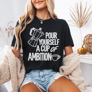 Puede incluir: Una camiseta negra de manga corta con un gr&aacute;fico blanco de una cafetera italiana y una taza de caf&eacute;, junto con el texto "POUR YOURSELF A CUP OF AMBITION" impreso en una fuente negrita y sin remates en el pecho.