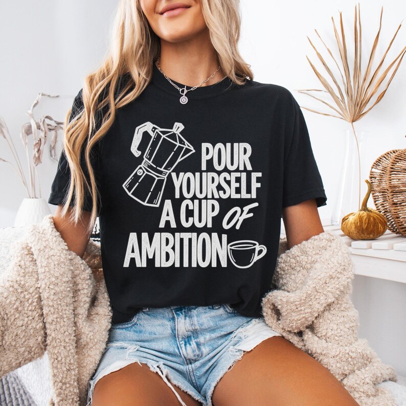 Puede incluir: Una camiseta negra de manga corta con un gr&aacute;fico blanco de una cafetera italiana y una taza de caf&eacute;, junto con el texto "POUR YOURSELF A CUP OF AMBITION" impreso en una fuente negrita y sin remates en el pecho.
