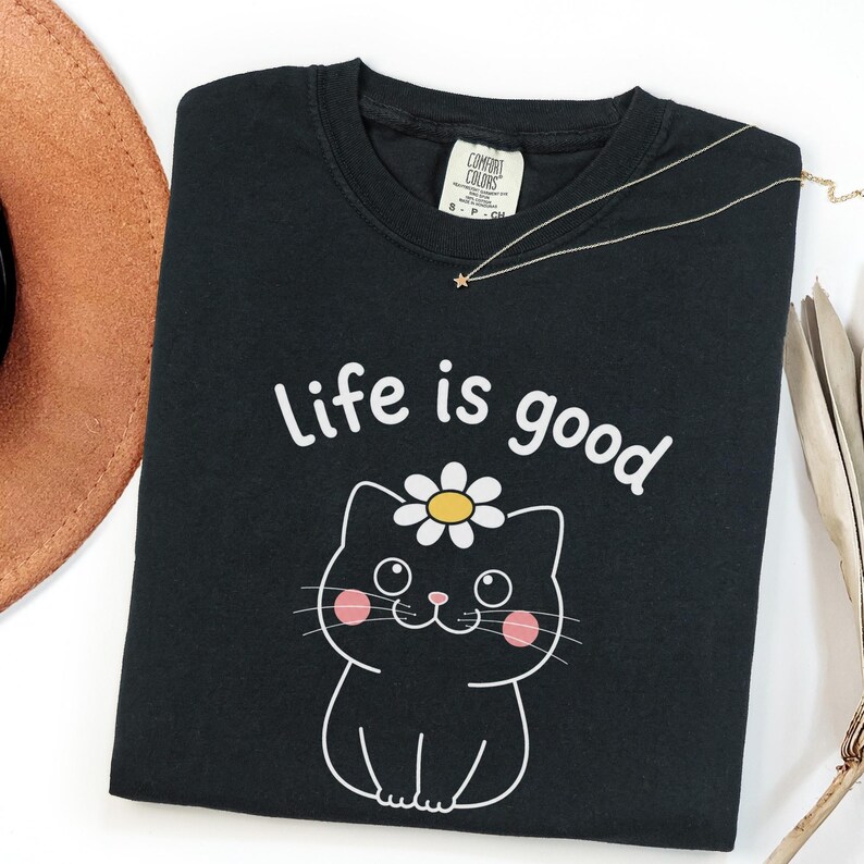 May include: Black t-shirt with the phrase "Life is good" and a cartoon cat wearing a daisy. A gold necklace with a star pendant and a brown hat are also visible.