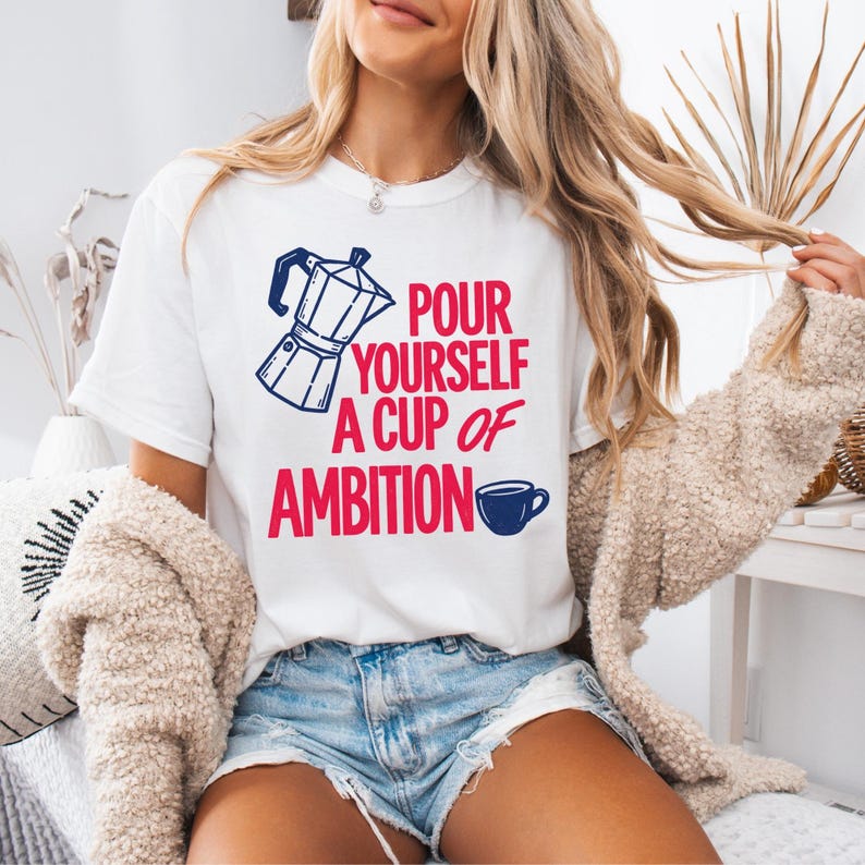 Puede incluir: Una camiseta blanca de manga corta con un gr&aacute;fico de una cafetera italiana azul y una peque&ntilde;a taza de caf&eacute; azul. El texto en rojo dice "POUR YOURSELF A CUP OF AMBITION". La camiseta se combina con pantalones cortos de mezclilla azul claro y un c&aacute;rdigan beige texturizado.