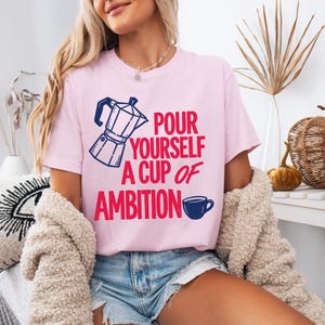 Puede incluir: Una camiseta de manga corta de color rosa claro con un gr&aacute;fico de cafetera moka en azul oscuro y el texto en rojo "POUR YOURSELF A CUP OF AMBITION" con un peque&ntilde;o icono de taza de caf&eacute; azul oscuro al final de la frase.