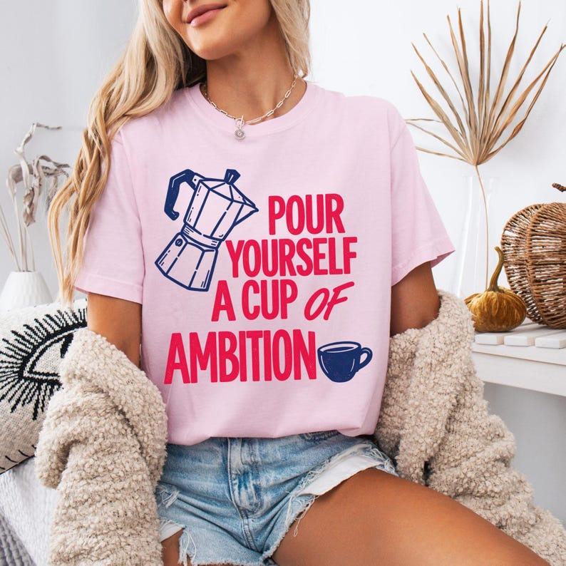 Puede incluir: Una camiseta de manga corta de color rosa claro con un gr&aacute;fico de cafetera moka en azul oscuro y el texto en rojo "POUR YOURSELF A CUP OF AMBITION" con un peque&ntilde;o icono de taza de caf&eacute; azul oscuro al final de la frase.