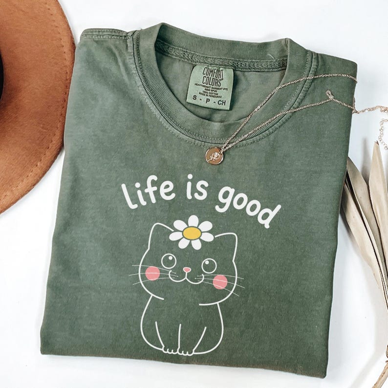May include: An olive green t-shirt with the phrase "Life is good" and a cartoon cat with a daisy on its head. A gold necklace is draped across the shirt. The shirt is labeled "Comfort Colors" and is a size small.