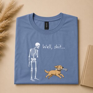 May include: A dusty blue t-shirt featuring a white graphic of a human skeleton standing next to a golden retriever dog. The dog is running and holding a human skeletal hand in its mouth. The text "Well, shit..." is printed in white above the dog.