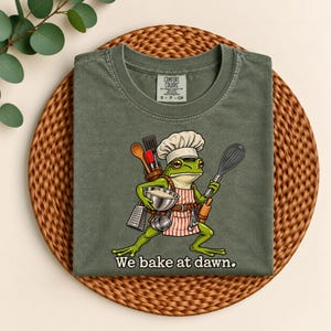 May include: A dark olive green t-shirt featuring a graphic of a frog wearing a chef hat and apron, holding a whisk and a bowl. The frog is equipped with various kitchen tools including a spatula and grater. The text "We bake at dawn." is printed below the graphic.