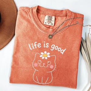 May include: Orange Comfort Colors t-shirt with the phrase "Life is good" and a cat graphic with a daisy. A silver necklace with a pendant is on the shirt. The shirt is folded and has a tag.