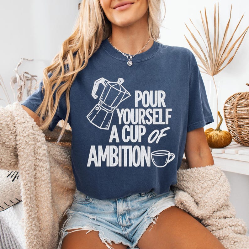 Puede incluir: Una camiseta azul marino de manga corta con un gr&aacute;fico blanco de una cafetera italiana y una taza de caf&eacute;, junto con el texto "POUR YOURSELF A CUP OF AMBITION" impreso en una fuente negrita y sin remates en el pecho de la prenda.