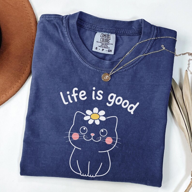 May include: A navy blue t-shirt with the phrase "Life is good" and a cartoon cat wearing a daisy. A gold necklace with a pendant is draped across the shirt. The shirt is folded and placed on a white surface.