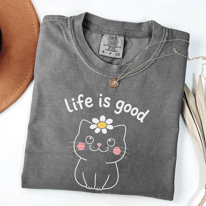 May include: A gray Comfort Colors t-shirt with the phrase "Life is good" and a cartoon cat with a daisy on its head. A silver necklace with a pendant and a brown hat are also visible.