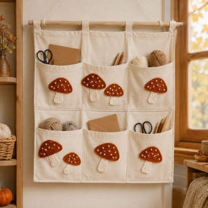 May include: A cream-colored canvas wall organizer with six pockets, each featuring a crocheted mushroom appliqué. The organizer hangs from a wooden rod and contains various craft supplies, including scissors, yarn balls, and pencils in the pockets.