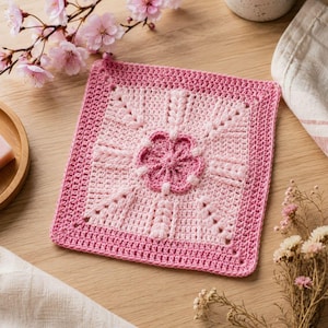 May include: A square, handmade crochet piece in shades of pink. The center features a flower design, surrounded by a lighter pink square, and a darker pink border. The piece is likely a coaster or decorative item.
