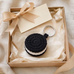 May include: A round, crocheted cookie keychain featuring two black textured circular pieces with a white center layer, attached to a silver metal ring. The item is presented inside a square cardboard gift box with a beige ribbon and tag.