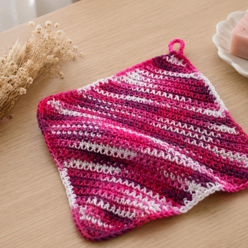 May include: A handmade crochet washcloth in pink, white, and purple. The washcloth has a diagonal striped design and a small loop for hanging. The item is square and looks to be made of soft yarn. The measurements are approximately 20 cm x 20 cm.