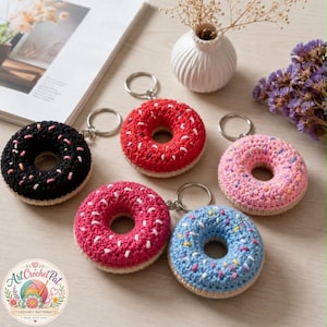Könnte beinhalten: Sechs gehäkelte Donut-Schlüsselanhänger in verschiedenen Farben: Schwarz, Rot, Rosa und Blau. Jeder Donut hat unterschiedlich farbige Streusel und einen silbernen Schlüsselring. Die Schlüsselanhänger sind auf einer hellen Oberfläche angeordnet. Durchmesser ca. 7 cm.