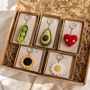 Puede incluir: Una colección de llaveros de ganchillo hechos a mano en una caja de regalo. Los llaveros presentan varios diseños, incluyendo una vaina de guisantes verdes, aguacate, corazón rojo, girasol y margarita. Cada llavero está empaquetado en una caja pequeña con una tarjeta que dice "handmade with love".