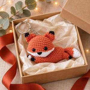 May include: A crocheted fox keychain featuring orange, white, and black yarn. The fox has black bead eyes, a black nose, and black-tipped ears and paws. A silver-toned metal split ring is attached to the top of the fox head with a short chain.