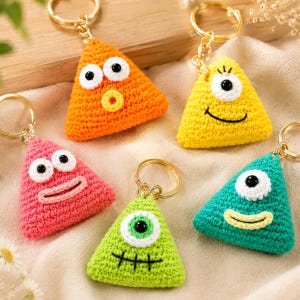 May include: Five triangular crochet keychains in orange, yellow, pink, lime green, and teal. Each keychain features a gold-tone metal ring and unique stitched facial expressions with white and black yarn eyes and embroidered mouths.