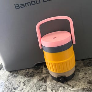May include: A multi-colored, stackable container with a pink handle and lid, a yellow mid-section, and a gray and tan base. The container is cylindrical and appears to be made of plastic. The text "Bambu Lab" is visible in the background.
