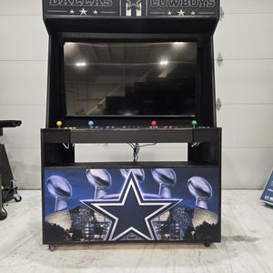 May include: A black arcade cabinet featuring Dallas Cowboys branding. The marquee displays the team name and star logo. The control panel has multi-colored joysticks and buttons. The base panel shows a large blue star and football trophies.