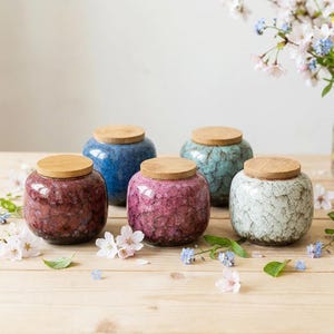 May include: Five ceramic jars with wooden lids in shades of blue, red, pink, and green sit on a wooden surface. The jars have a textured, crackled finish. Cherry blossoms and other flowers are scattered around the jars.