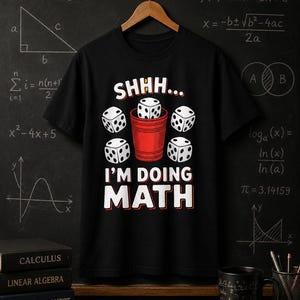 Op de afbeelding: Een zwart T-shirt met korte mouwen en een grafische print van een rode plastic beker gevuld met witte dobbelstenen, omringd door vier extra dobbelstenen. De tekst luidt SHHH... I'M DOING MATH in witte hoofdletters met een rode rand.