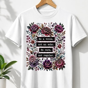 May include: A white short-sleeve t-shirt featuring a central rectangular graphic surrounded by dark purple, pink, and green floral illustrations. The text reads: Be a voice, not an echo. Be rare, not regular. The shirt is displayed on a wooden hanger.