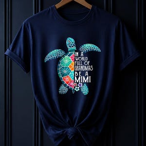 May include: A navy blue short-sleeve t-shirt featuring a graphic of a sea turtle with a colorful mosaic shell. The text on the shirt reads, "IN A WORLD FULL OF GRANDMAS BE A MIMI," accompanied by small floral accents near the turtle's lower body.