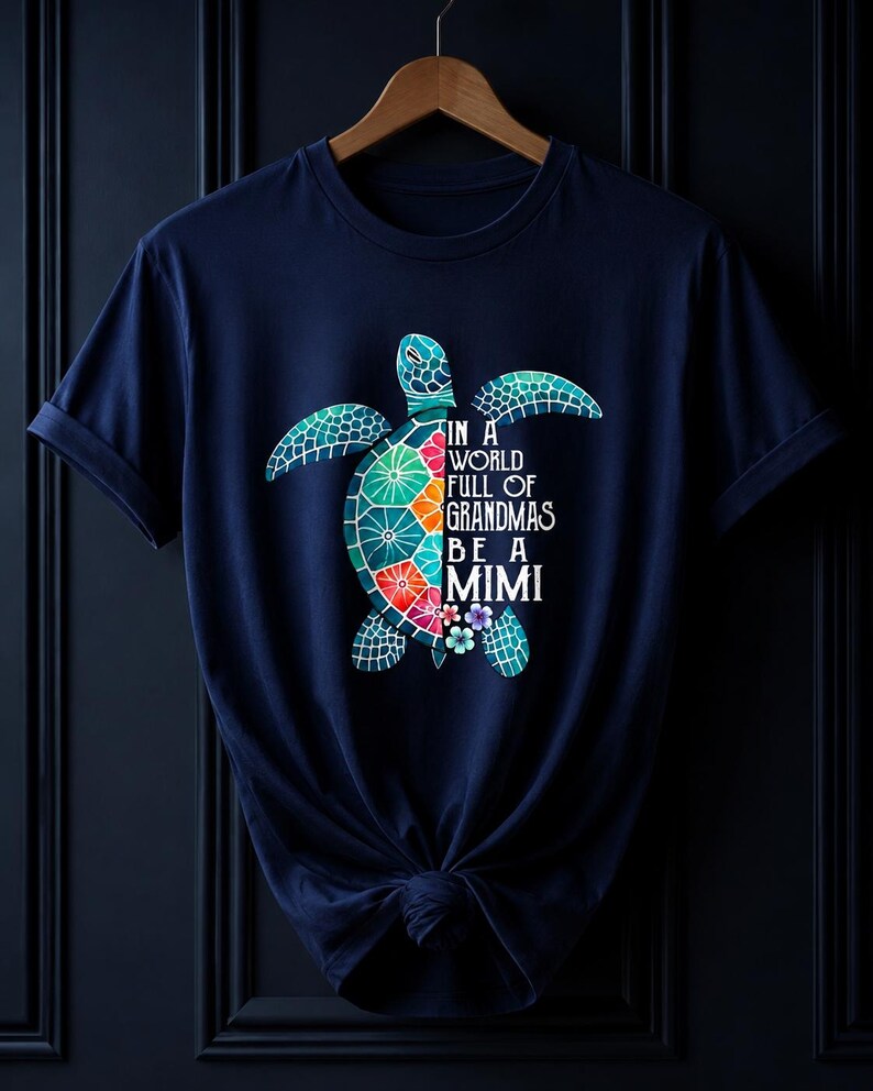 May include: A navy blue short-sleeve t-shirt featuring a graphic of a sea turtle with a colorful mosaic shell. The text on the shirt reads, "IN A WORLD FULL OF GRANDMAS BE A MIMI," accompanied by small floral accents near the turtle's lower body.