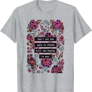 May include: A heather gray short-sleeve t-shirt featuring a central graphic of dark pink and purple flowers and leaves. The text "Don't let the ugly in others kill the beauty in you." is printed in white inside black rectangular boxes.