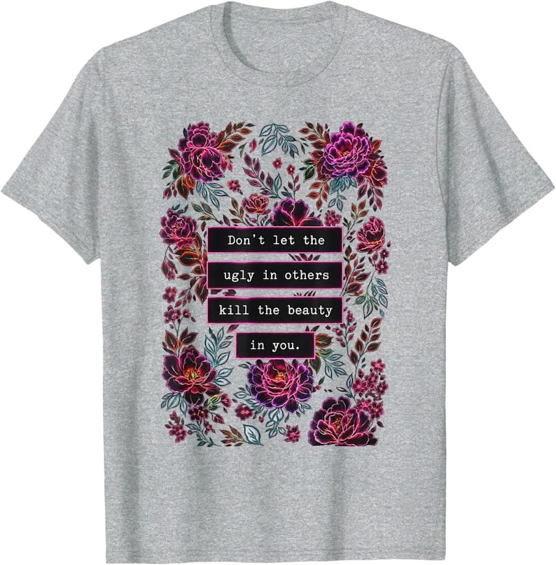 May include: A heather gray short-sleeve t-shirt featuring a central graphic of dark pink and purple flowers and leaves. The text "Don't let the ugly in others kill the beauty in you." is printed in white inside black rectangular boxes.