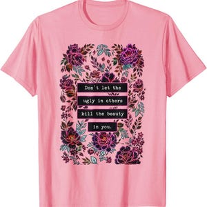 May include: A light pink short-sleeved t-shirt featuring a central graphic of dark purple and magenta flowers with green leaves. The text "Don't let the ugly in others kill the beauty in you." is printed in white on black rectangular bars.