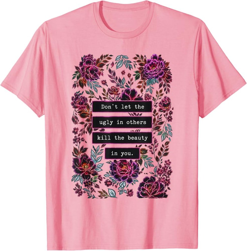 May include: A light pink short-sleeved t-shirt featuring a central graphic of dark purple and magenta flowers with green leaves. The text "Don't let the ugly in others kill the beauty in you." is printed in white on black rectangular bars.