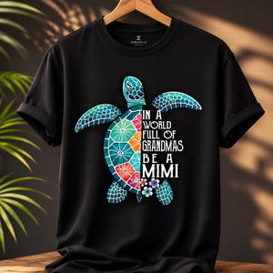 May include: A black short-sleeved t-shirt featuring a graphic of a sea turtle with a colorful, patterned shell on the left side and the white text "IN A WORLD FULL OF GRANDMAS BE A MIMI" on the right side, accented with small floral details.