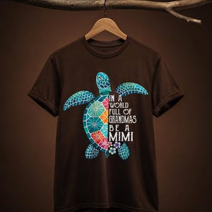 May include: A dark brown short-sleeve t-shirt featuring a colorful sea turtle graphic on the front. The turtle shell is divided into vibrant segments of blue, green, orange, and red. The text reads: IN A WORLD FULL OF GRANDMAS BE A MIMI.