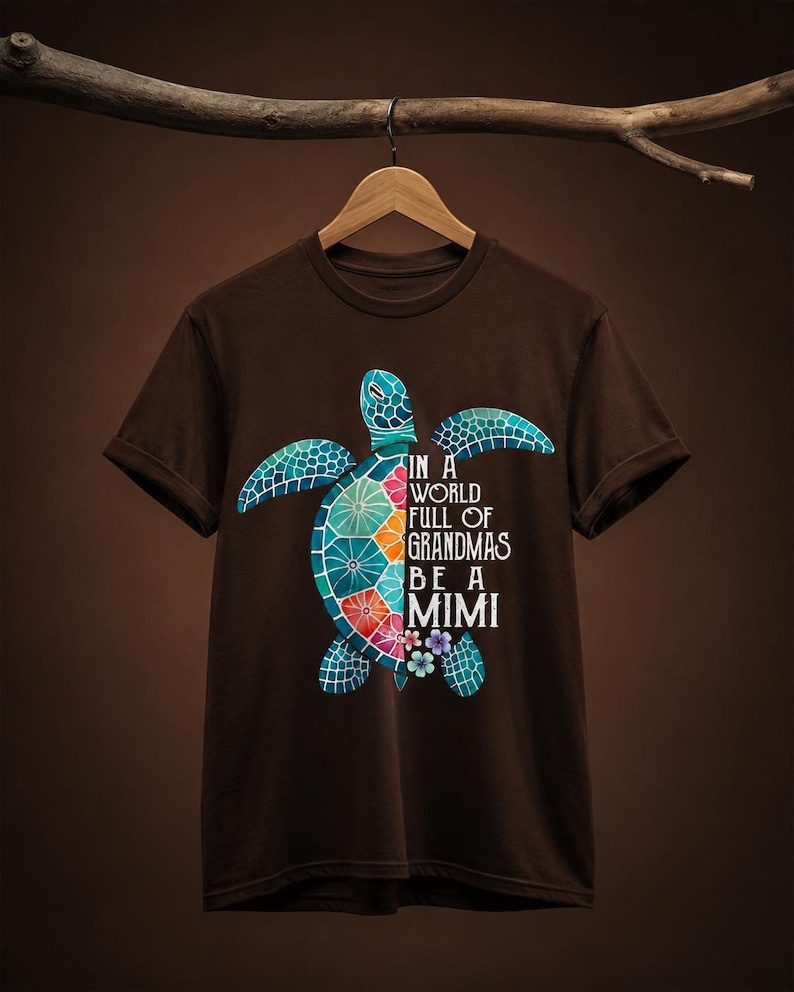 May include: A dark brown short-sleeve t-shirt featuring a colorful sea turtle graphic on the front. The turtle shell is divided into vibrant segments of blue, green, orange, and red. The text reads: IN A WORLD FULL OF GRANDMAS BE A MIMI.