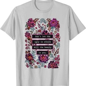 May include: A light gray short-sleeve t-shirt featuring a central rectangular graphic of dark purple and pink flowers and leaves. The text "Don't let the ugly in others kill the beauty in you." is printed in white inside black rectangular bars.