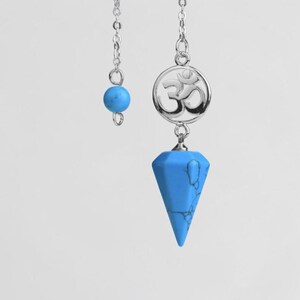 Puede incluir: Un péndulo plateado con una punta de piedra turquesa y un dije plateado con el símbolo Om. Una pequeña cuenta turquesa cuelga de una cadena separada. El péndulo está diseñado para prácticas espirituales y adivinación.