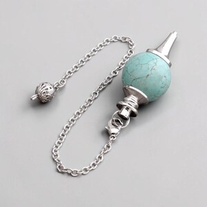 May include: A pendulum with a turquoise stone sphere and a silver-colored chain. The sphere has a cracked pattern and is attached to a silver metal cone and a chain with a decorative ball at the end. The chain is connected to the sphere.