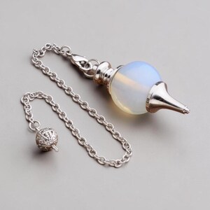 May include: A pendulum with a silver chain and a teardrop-shaped, opalescent crystal. The pendulum has a silver cap and a pointed tip. A smaller, decorative silver ball hangs from the chain.