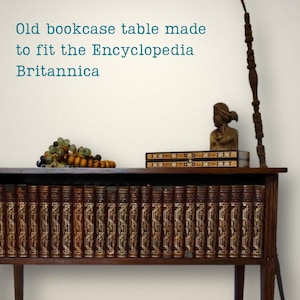 May include: A dark wood bookcase table filled with a row of vintage books with gold-embossed spines. On top sits a decorative rectangular box, a small brown bust, and a cluster of glass grapes. Text reads: Old bookcase table made to fit the Encyclopedia Britannica.