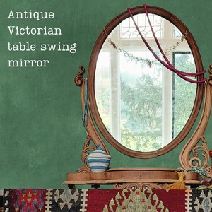 May include: An oval wooden table swing mirror with ornate carved supports. The text "Antique Victorian table swing mirror" is visible on the left. A small blue and white striped lidded container sits on the base, with a red cord draped over the glass.
