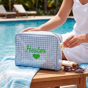 May include: A rectangular light blue and white gingham toiletry bag featuring the name Hester embroidered in bright green script, accompanied by a green heart icon. The bag has a zipper closure and a structured, boxy shape.