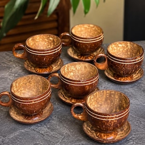以下が含まれることがあります： ココナッツの殻で作られた手作りのカップとソーサー6客セット。茶色のカップは自然な質感で、白いバンドがアクセントになっています。各カップには取っ手があり、お揃いのソーサーの上に置かれています。セットは濃い灰色の表面に配置されています。